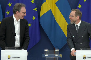 Två personer står vid var sin talarpulpet halv vända mot varandra och ler vänligt mot varandra.