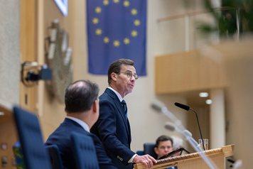 Ulf Kristersson står i en talarstol i riksdagen. I bakgrunden syns talman Andreas Norlén och EU:s flagga.