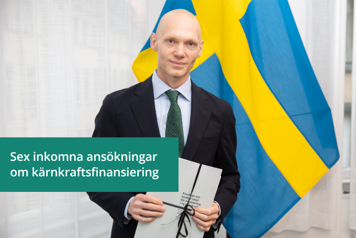 Finansmarknadsminister står framför en svensk flagga och håller i en ansökan i en pappersmapp.