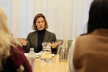 Jämställdhetsminister Nina Larsson sitter vid ett bord och pratar. I förgrunden syns två personer bakifrån.