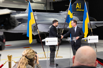 Volodymyr Zelensky och Ulf Kristersson står på en pressträff och skakar hand. I bakgrunden står ett JAS Gripen samt ukrainska och svenska flaggor. I förgrunden syns journalister.