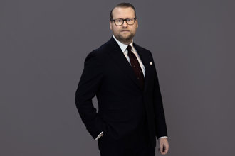Statssekreterare Simon Westberg står framför en grå bakgrund, klädd i mörk kostym, vit skjorta och mörk slips. Han bär glasögon och ser rakt mot kameran med ena handen i fickan.