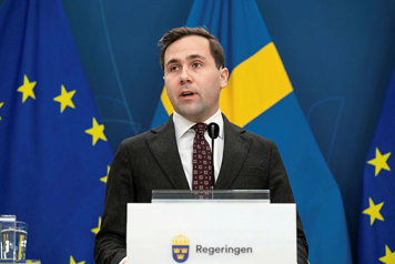 Statsrådet avbildad i halvfigur bakom en talarpupet. Flaggor i fonden.