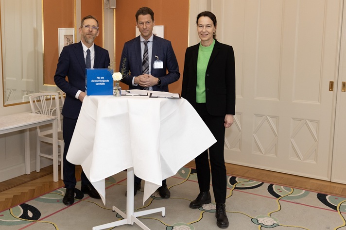 Tre personer står kring ett högt bord med dokument och en blå pärm i ett mötesrum.