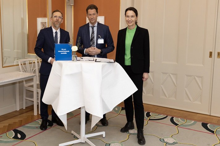 Tre personer står kring ett högt bord med dokument och en blå pärm i ett mötesrum.