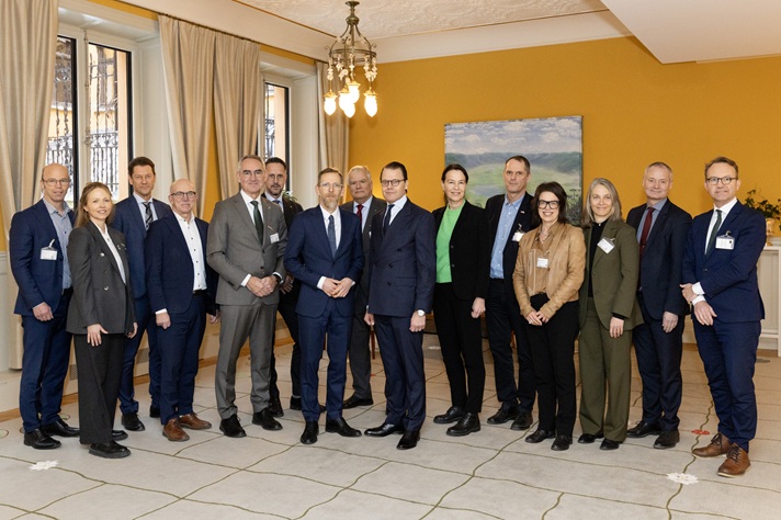 En grupp på femton personer står samlade i ett mötesrum med gula väggar och stora fönster. De är formellt klädda och står uppställda i en halvcirkel för ett gruppfoto.