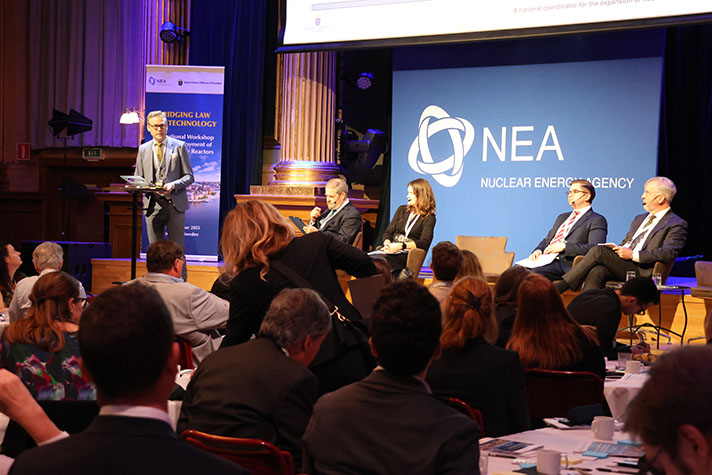 En man står upp på scenen och talar, tre andra personer sitter intill.