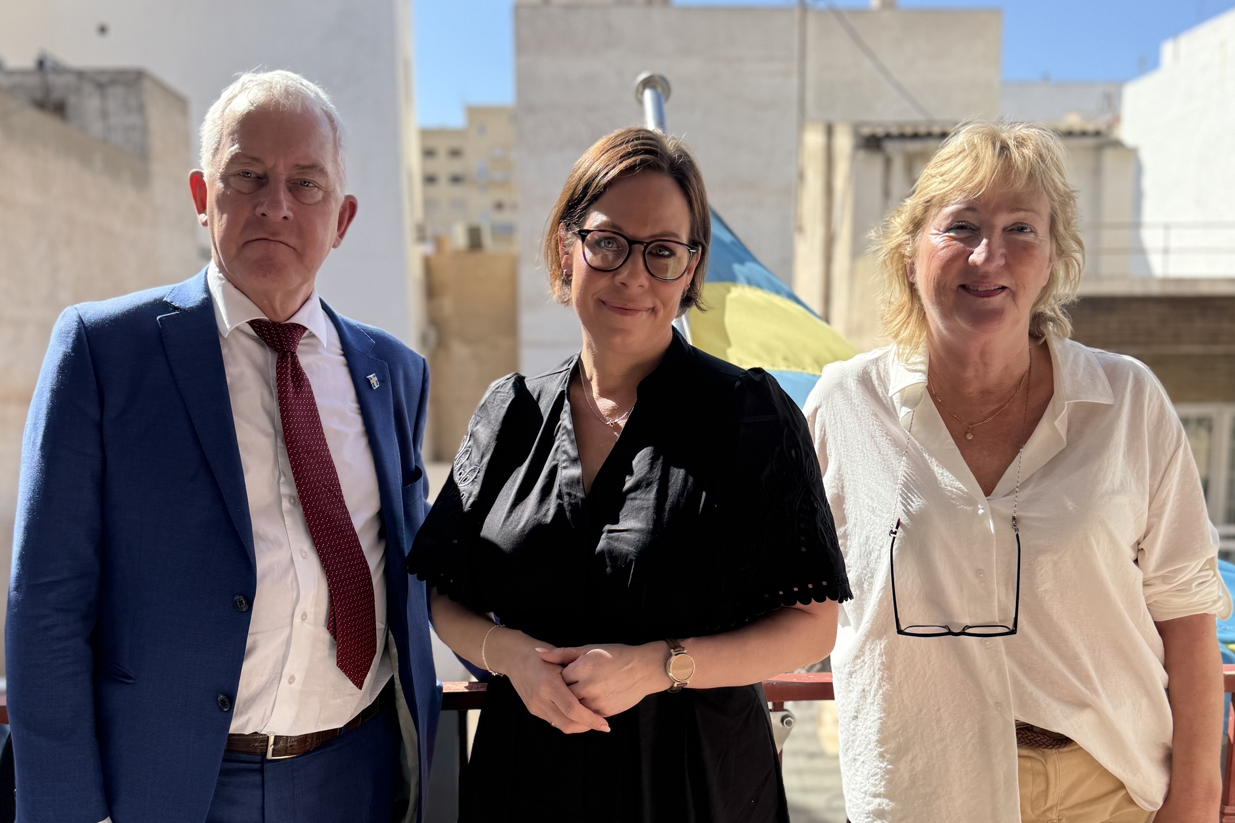 Utrikesministern, honorärkonsul Björn Sandström och kanslist Caroline Lamenoise står utomhus framför en svensk flagga.