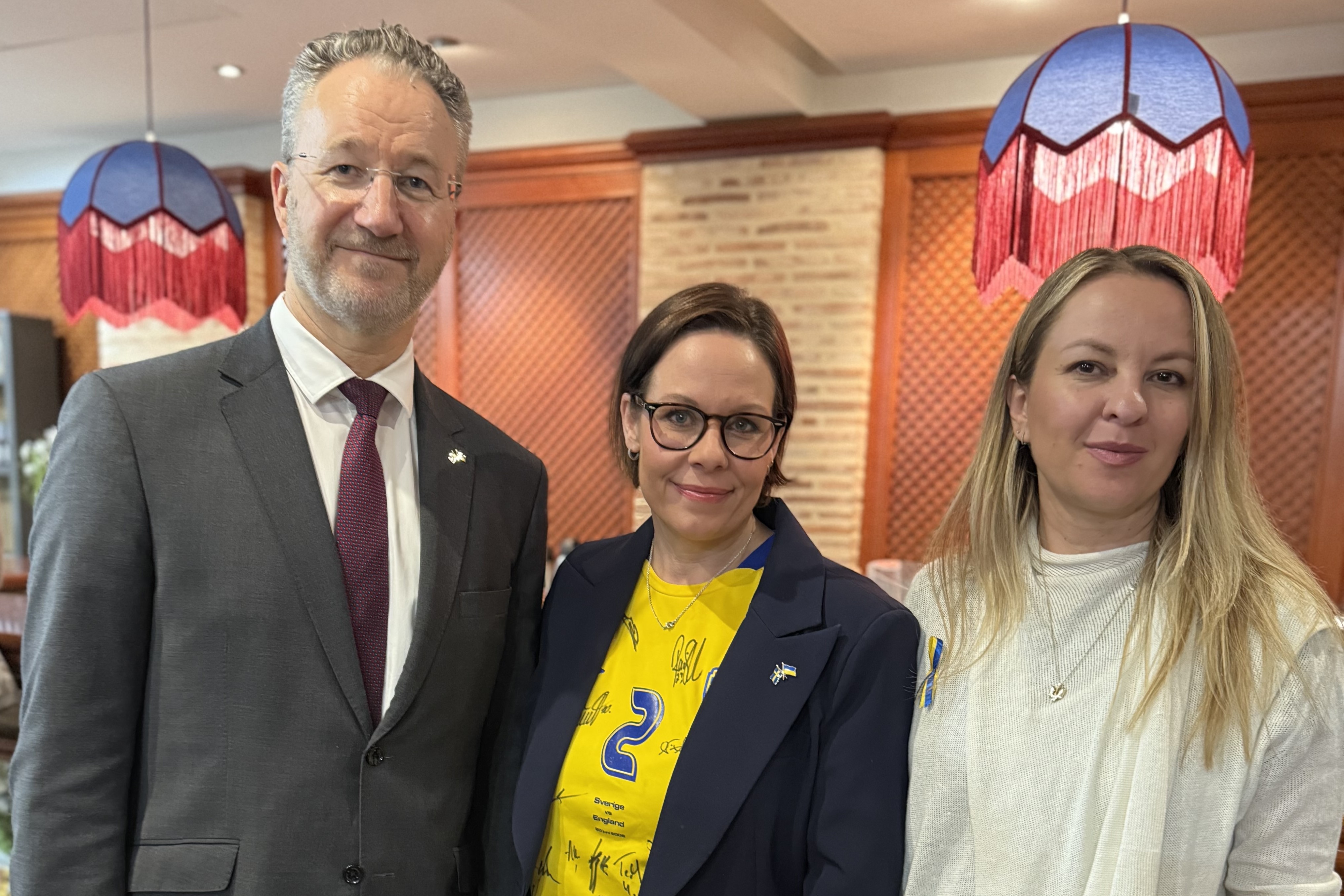 Utrikesministern, Sveriges ambassadör i Spanien och Ukrainas ambassadör i Spanien står bredvid varandra och ler in i kameran. Utrikesministern har på sig en svensk landslagströja.