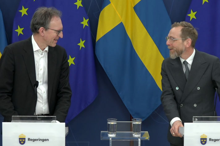 Två personer står vid var sin talarpulpet halv vända mot varandra och ler vänligt mot varandra.