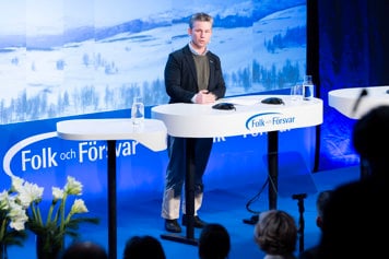 Pål Jonson står på en scen vid ett ståbord och talar. I bakgrunden syns ett vinterlandskap på stora skärmar.