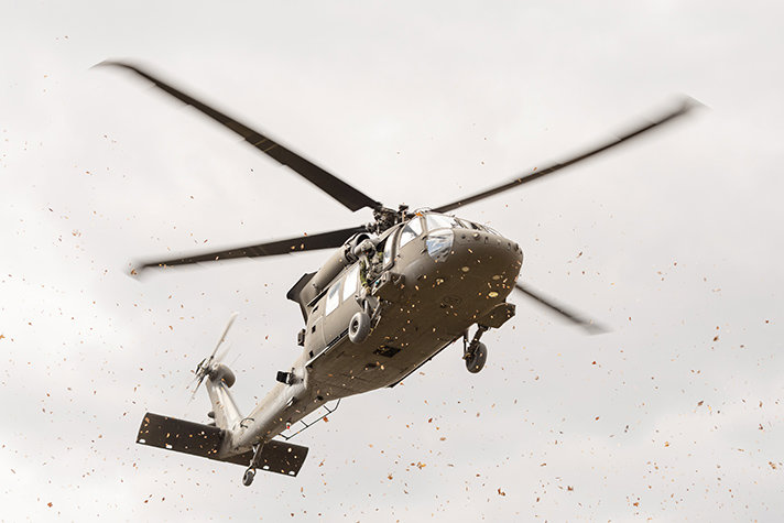 En helikopter 16 (Blackhawk) på väg för landning. En stor mängd löv flyger runt i vinden av rotorbladen. En besättningsmedlem sitter halvvägs utanför helikoptern och tittar ned.