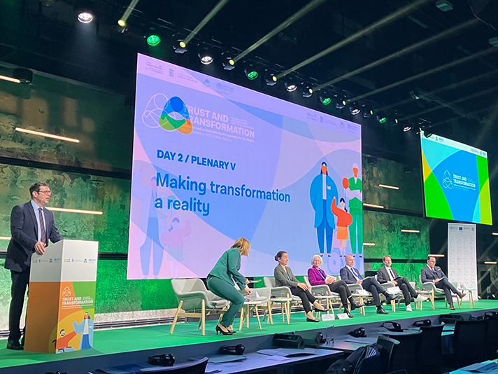Foto på ett panelsamtal på en scen. En stor skärm bakom deltagarna med texten "Making transformation a reality" 