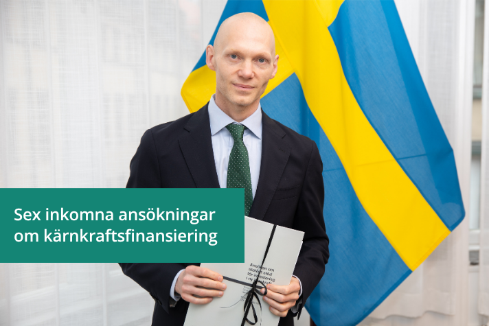 Finansmarknadsminister står framför en svensk flagga och håller i en ansökan i en pappersmapp.