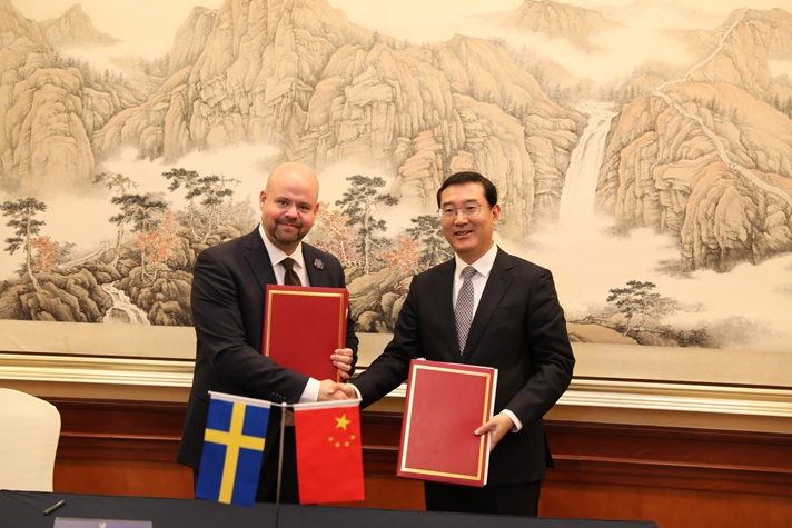 Två män i kostym skakar hand och håller i varsin pärm. I förgrunden visas svenska och kinesiska flaggan.
