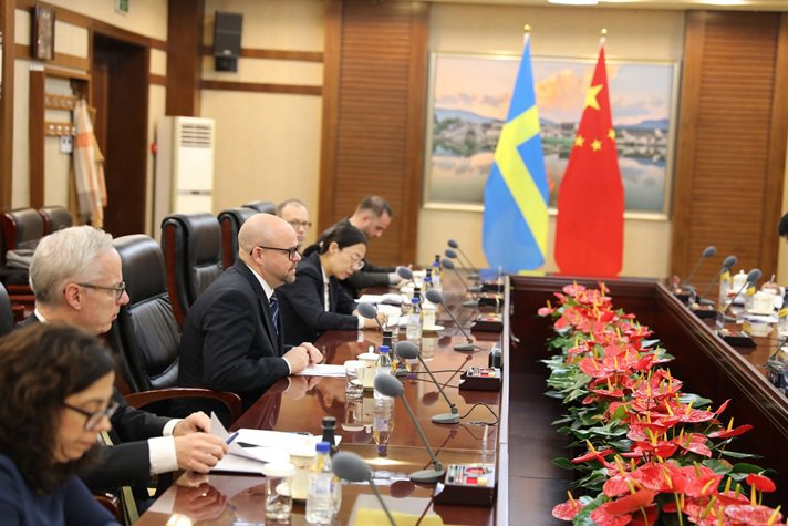 En grupp personer sitter vid ett bord och bläddrar i papper. I bakgrunden visas svenska och kinesiska flaggan.