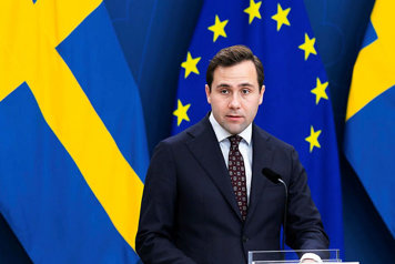 Bistånds- och utrikeshandelsminister Benjamin Dousa står vid en talarpulpet, det är flaggor i fonden.