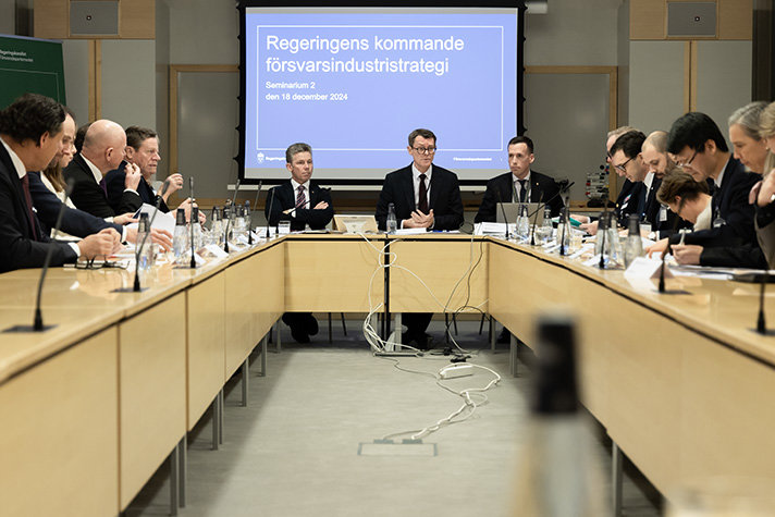 Pål Jonson sitter med statssekreterare Peter Sandwall som hälsar personerna i rummet välkomna till seminariet. Personerna sitter vid egna bord runt ett öppet utrymme med statssekreteraren och försvarsministern vid kortändan.