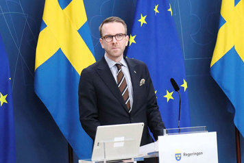 Infrastruktur- och bostadsminister Andreas Carlson fotograferad i halvfigur vid en talarpulpet med svenska flaggor i bakgrunden.