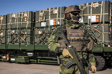 En soldat i uniform står framför en materieltransport.