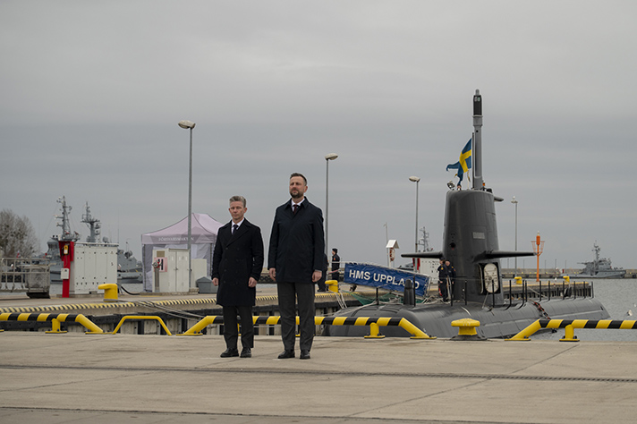 Pål Jonson och Władysław Kosiniak-Kamysz står på en kaj i Gdynia. Det är mulet och i bakgrunden syns en svensk ubåt ligga förtöjd.