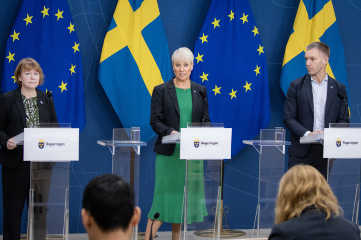 Danielsson, Tenje och Slottner står bakom varsin talarstol rí regeringens pressrum, framför svenska och EU-flaggor.