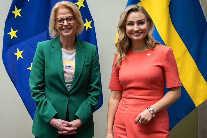 Elisabeth Svantesson och Ebba Busch står sida vid sida och ler mot kameran, framför EU-flaggor och svenska flaggor. Svantesson bär grön kavaj och Busch en korallfärgad klänning.