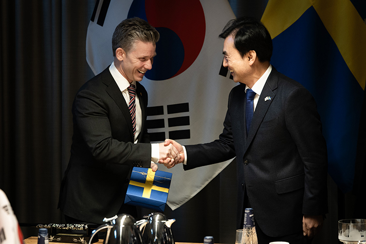 Pål Jonson skakar hand med Ahn Gyu-back efter denne har överlämnat en gåva inslaget i papper med den svenska flaggan. I bakgrunden syns den Sydkoreanska flaggan.