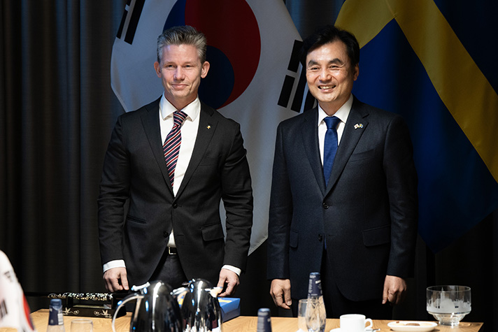 Pål Jonson och Ahn Gyu-back står sida vid sida vända mot kamerorna som inte syns i bild. I bakgrunden syns Sydkoreas och den svenska flaggan.