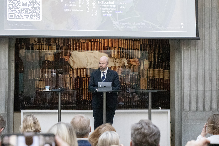 En man i mörk kostym står på en scen och pratar framför en publik