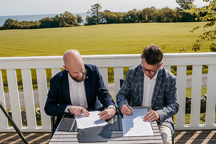 På en solig terrass sitter Carl-Oskar Bohlin och Torsten Schack Pedersen vid ett bord sida vid sida och undertecknar avsiktsförklaringen. 