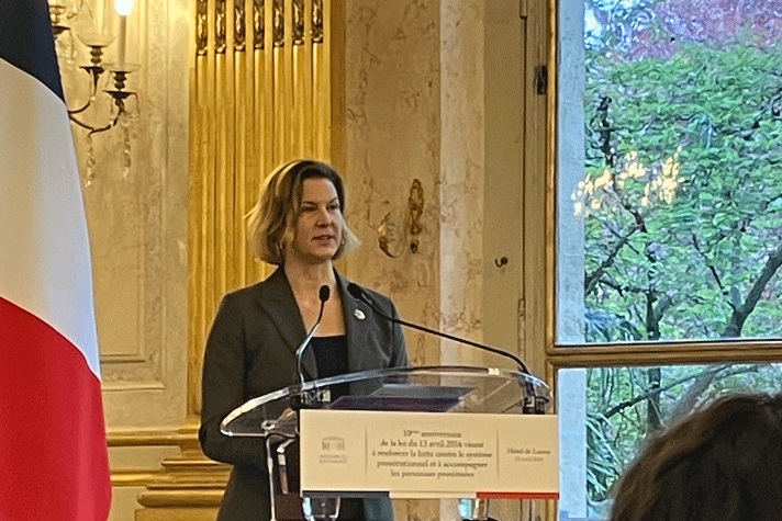 Nina Larsson står bakom en talarstol. Till vänster syns den franska flaggan och till höger ett fönster med träd utanför. I bakgrunden syns guldprydda pelare och en marmorvägg.