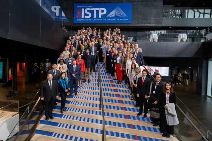En stor grupp delegater från olika länder står på en bred trappa under en banderoll där det står "ISTP 2025: International Summit on the Teaching Profession".