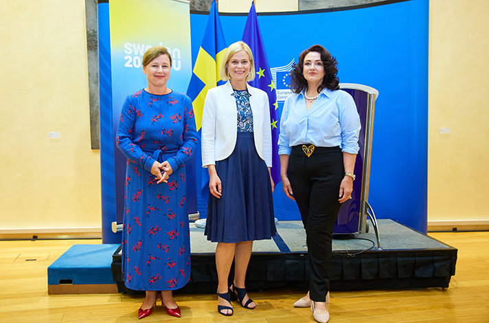 Vĕra Jourová, Paulina Brandberg och Helena Dalli står bredvid varandra och ler in i kameran. I bakgrunden syns en talarstol och flaggor.