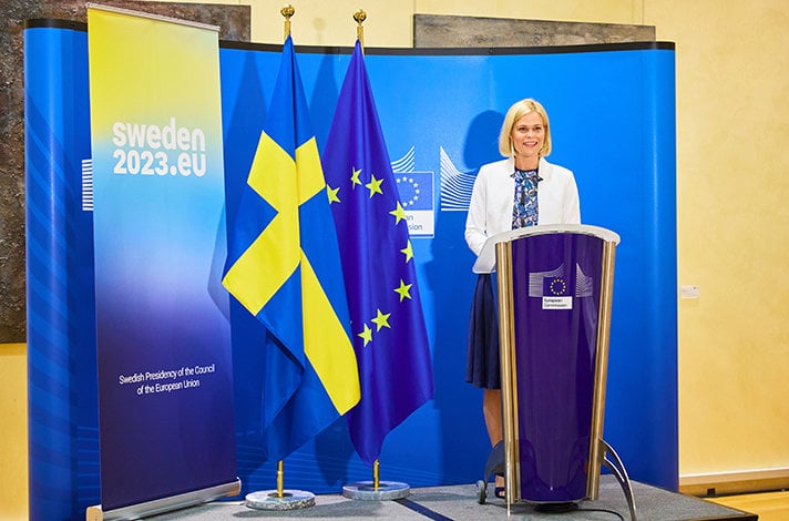 Paulina Brandberg står i en talarstol. Bredvid henne syns en EU-flagga och en flagga för det svenska ordförandeskapet i EU:s ministerråd.