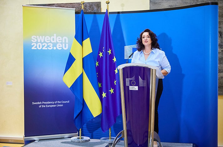 Helena Dalli står bakom en talarstol och pratar. I bakgrunden syns en EU-flagga och en flagga för det svenska ordförandeskapet i EU:s ministerråd.