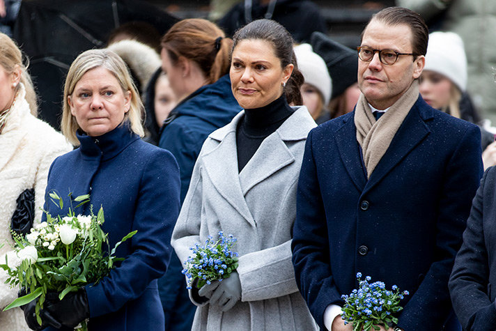 Andersson och kronprinsessparet står utomhus med blommor i händerna under en minnesceremoni