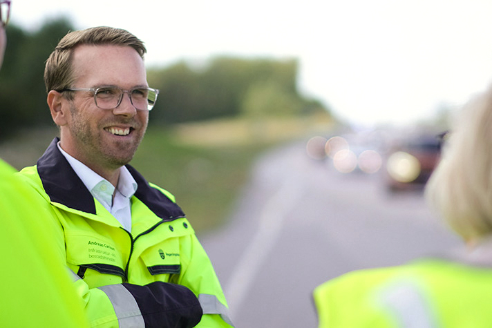 Utomhusbild på Carlson som bär en varseljacka i neongrönt och står vänd mot kameran. Axlarna från två andra personer i varselklädsel syns också. I bakgrunden ett sommarlandskap med en landsväg trafikerad med ett flertal bilar, bakgrunden är mycket oskarp.