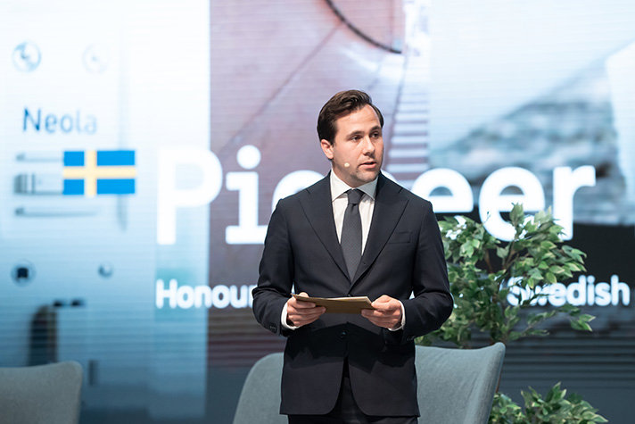 Inomhusbild med Dousa, iförd headset, står framför en bakgrund med svensk flagga och texten Pioneer.