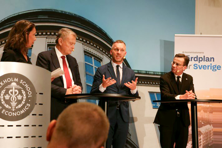 Inomhusbild. Slottner står i mitten av bilden vid ett pelarbord vänd mot kameran. Till vänster om Slottner står två personer, och till höger står statsministern vid ett annat pelarbord. Alla bär headset. I vänster hörn syns ett märke med texten Stockholm School of Economics. Till höger i bild en roll-up med texten ”AI-kommissionens färdplan för Sverige.”