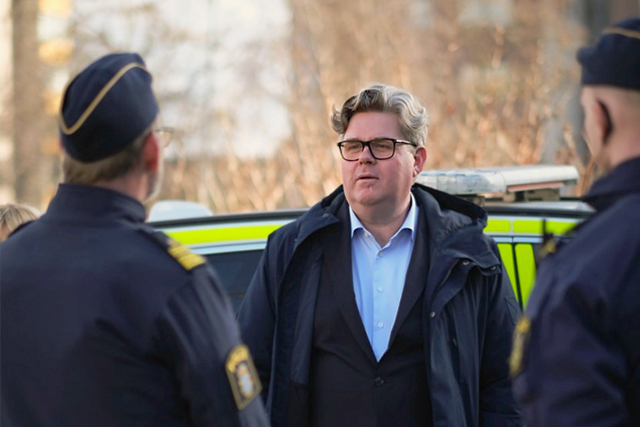 Utomhusbild med Strömmer i helfigur vänd mot kameran med en polisbil bakom sig. Framför honom, till höger och till vänster, står två poliser med ryggen mot kameran och i samtal med Strömmer. Bakgrunden är oskarp och man anar ett flerfamiljshus samt en genombruten växtlighet.