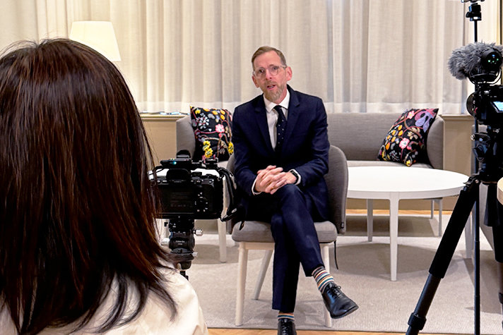 Inomhusbild med Forssmed i en intervjusituation. Han sitter i mitten av bilden i en klädd stol vänd mot kameran. Framför honom till vänster en person vars bakhuvud syns och som har en kamera på stativ framför sig vänd mot Forssmed. Till höger i bild syns ytterligare en kamera på stativ och med en lurvig mikrofon. I bakgrunden en neutral soffa med två blommönstrade kuddar och ett lågt soffbord. Samt hängande gardiner som täcker hela bakre