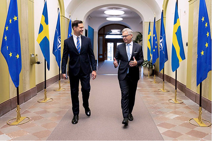 Inomhusbild på Forssell och Brunner som går mot kameran, i en korridor med flaggor längs sidorna, växelvis svenska, Nato- och EU-flaggor.