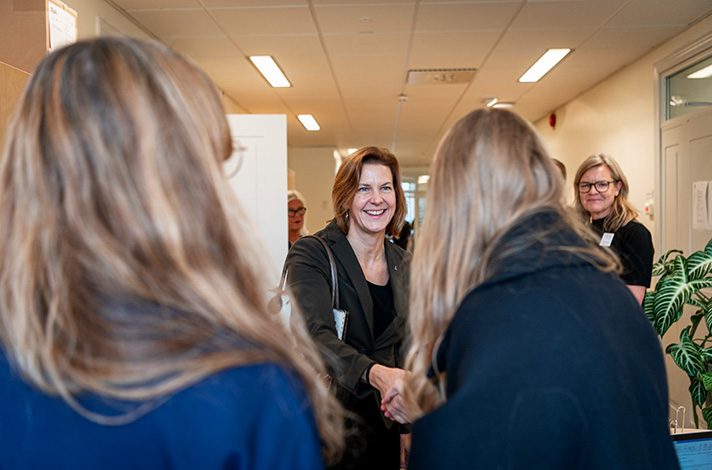 Inomhusbild i kontorsmiljö. Larsson vänd mot kameran skakar hand med en person till höger i bild som har ryggen mot kameran. Ytterligare en person står med ryggen mot kameran till vänster i bild. Snett bakom Larsson syns flera andra personer som tittar mot de som hälsar.