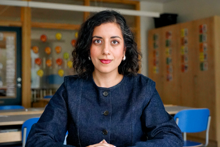 Porträttbild på Mohamsson i halvfigur, inomhus i en skolmiljö. Hon sitter och ser rakt mot kameran. I bakgrunden syns ett klassrum med skåp, bänkar och stolar.