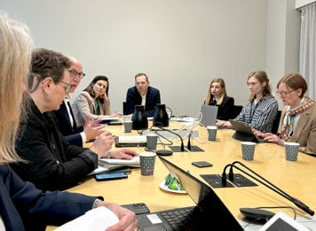 En grupp personer sitter runt ett konferensbord och lyssnar uppmärksamt på en som talar. Datorer och kaffemuggar framför står framför dem.