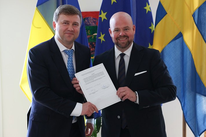 Två leende män i kostym håller i ett papper.