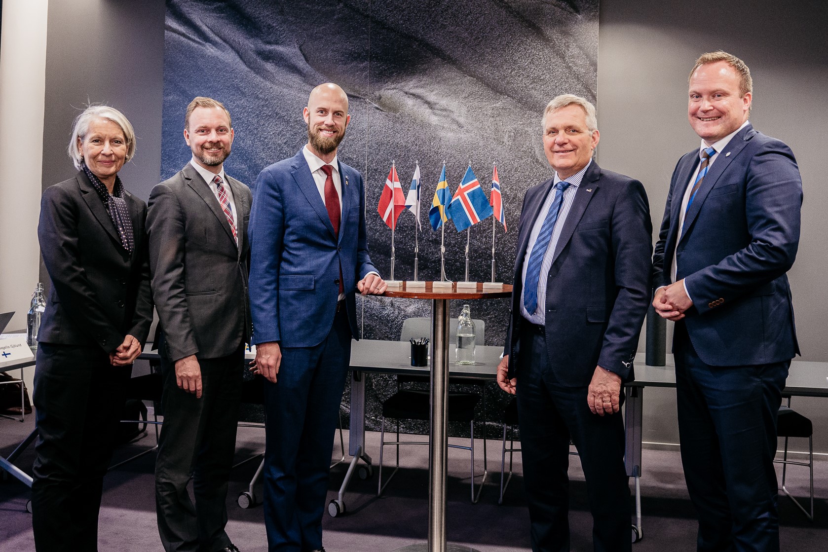 Representanter från respektive deltagande nation står bredvid varandra och ler. I mitten står ett bord med respektive lands flaggor.