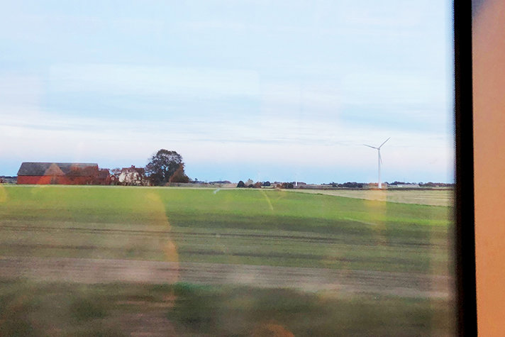 Foto från ett tågfönster som visar ett landskap med ett vindkraftverk i bakgrunden