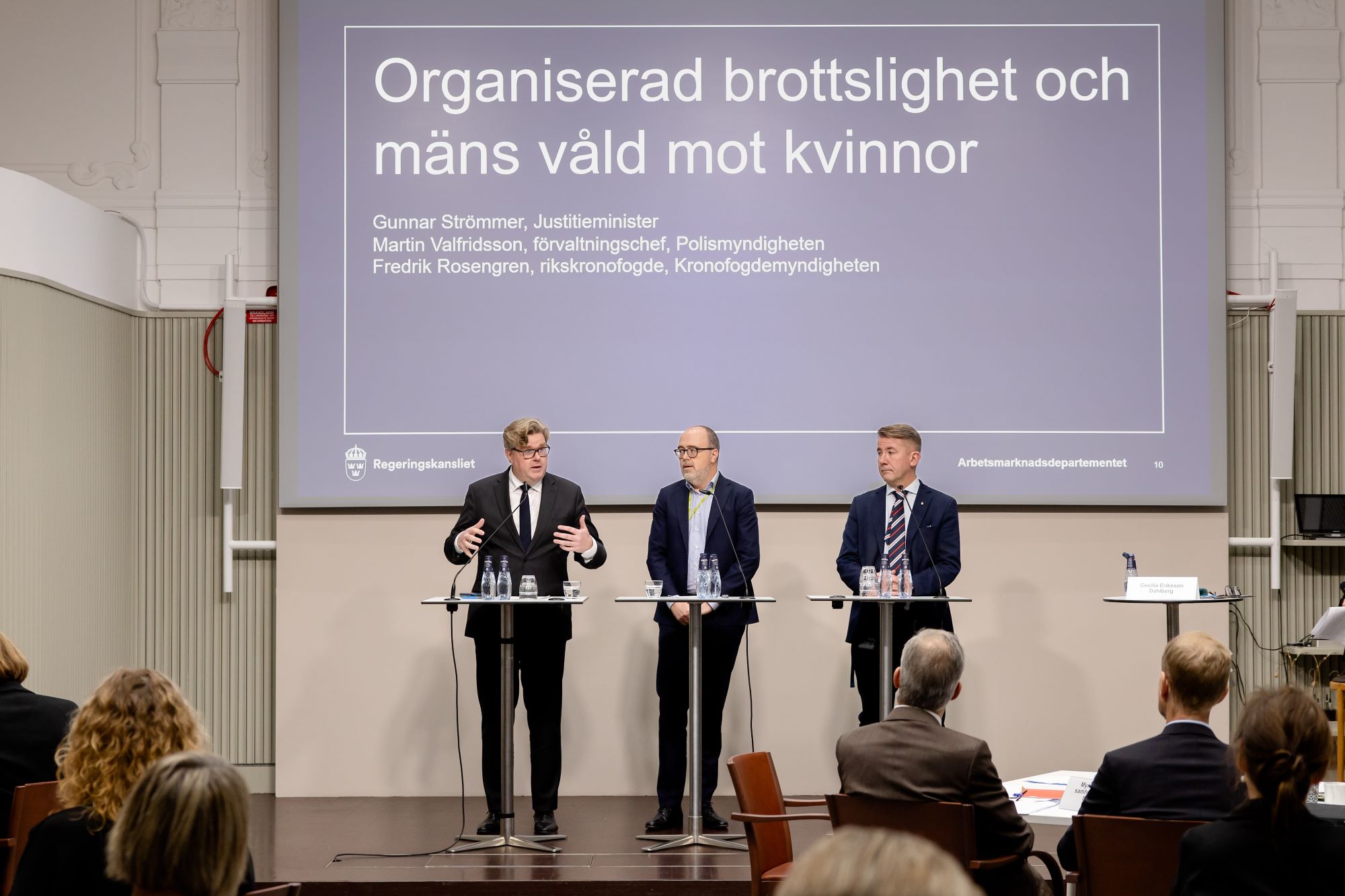 Tre personer står och pratar på en scen. I bakgrunden syns en presenetationsbild.